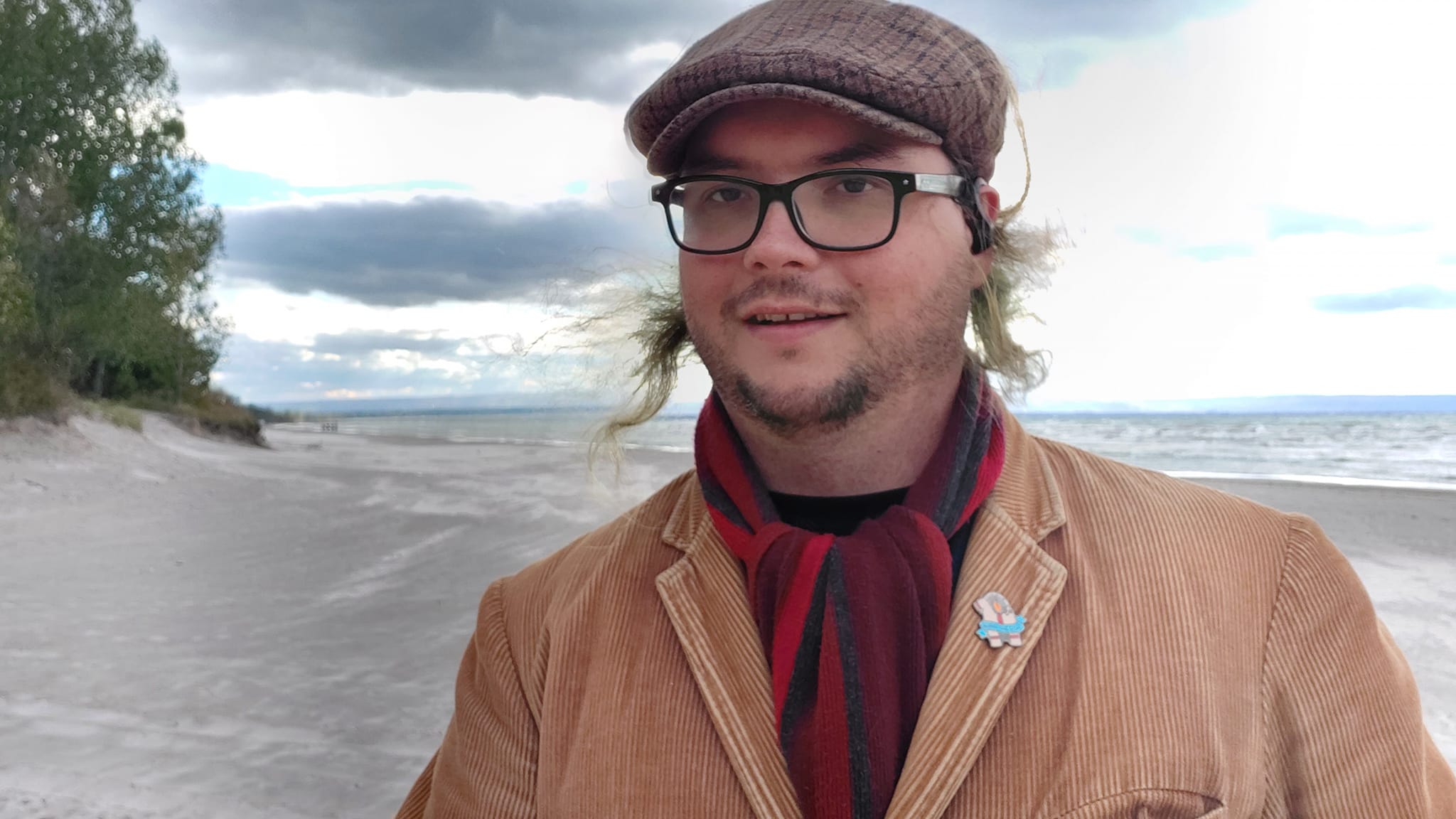 A man with long green hair stands on a beach, big water behind him